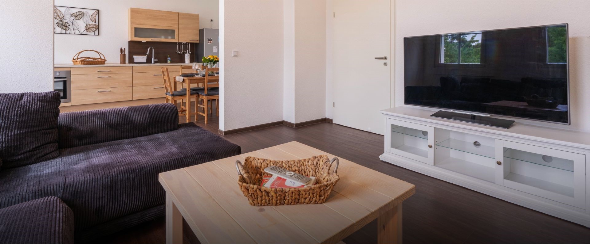 Modernes Wohnzimmer mit offener Küche, einem Fernseher, dunkelgrauem Sofa und hellem Holzcouchtisch